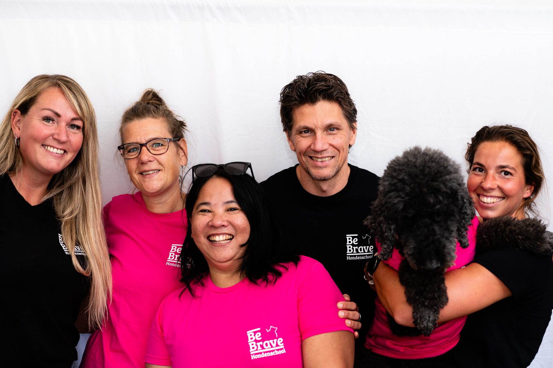 Het team van Be Brave in roze en zwarte shirts, lachend naast elkaar met een zwarte krulhond erbij